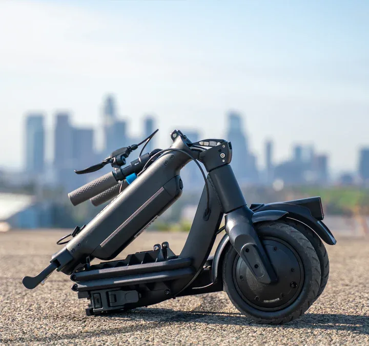 Riley RS3 electric scooter on outdoor surface with city skyline background, showing handlebars, motor, and pneumatic tyres