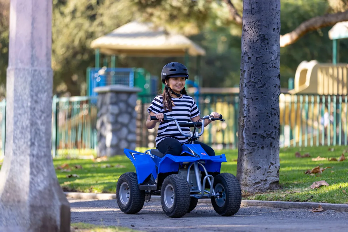 Razor Dirt Quad 24V electric ride-on quad in blue ridden by a child wearing a helmet on a park path, showing knobby tyres and steel frame