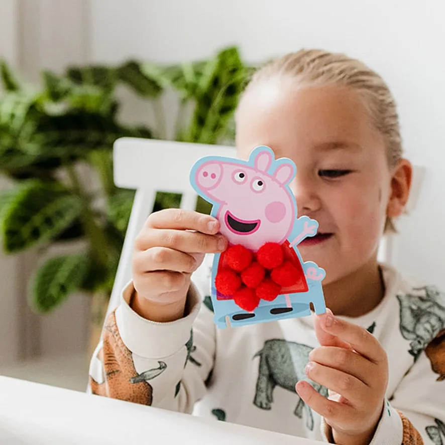 Young child holding a completed Peppa Pig pompom card decorated with red fluffy pom-poms covering the character's body