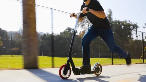 A teenager riding the Razor Carbon Lux black kick scooter outdoors on a paved path in bright sunshine, showing the scooter in motion.