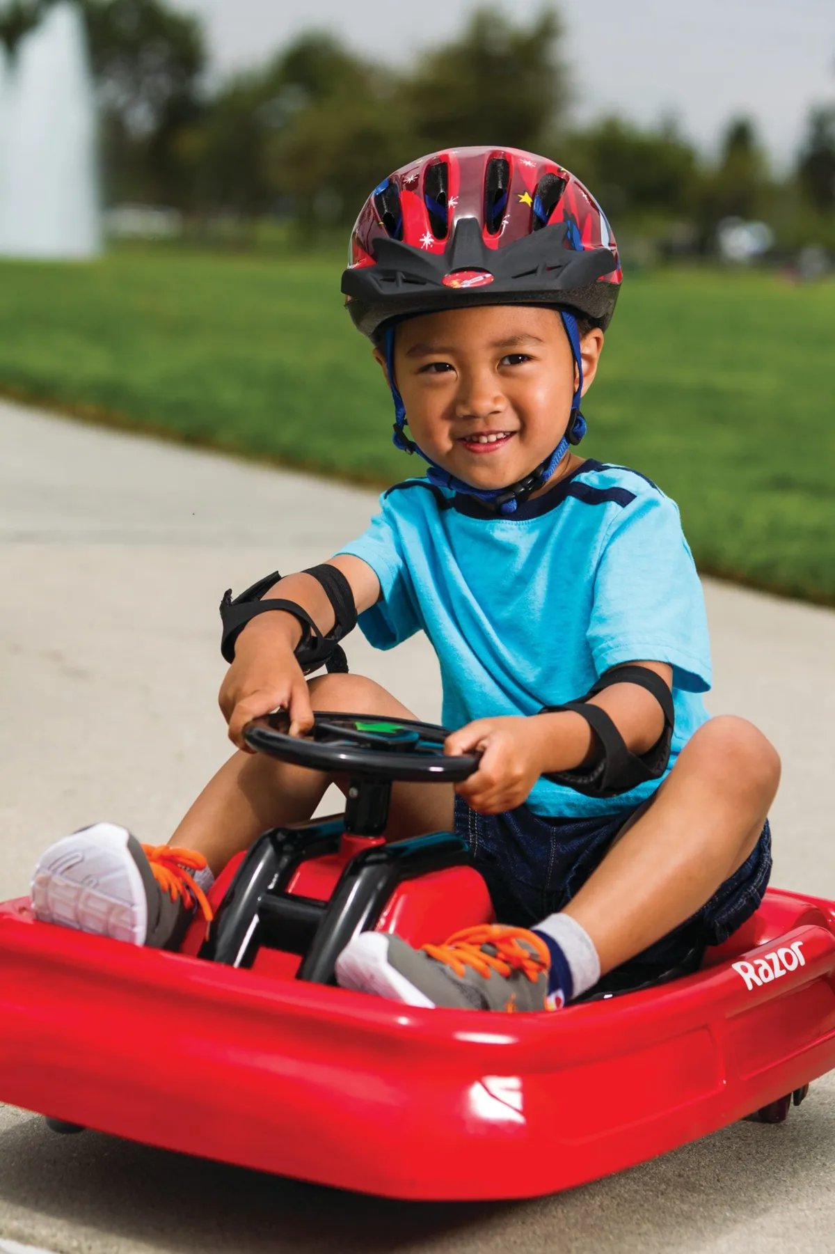 Young boy wearing a helmet and pads riding the red Razor Lil Crazy electric drift cart on a driveway, smiling at camera