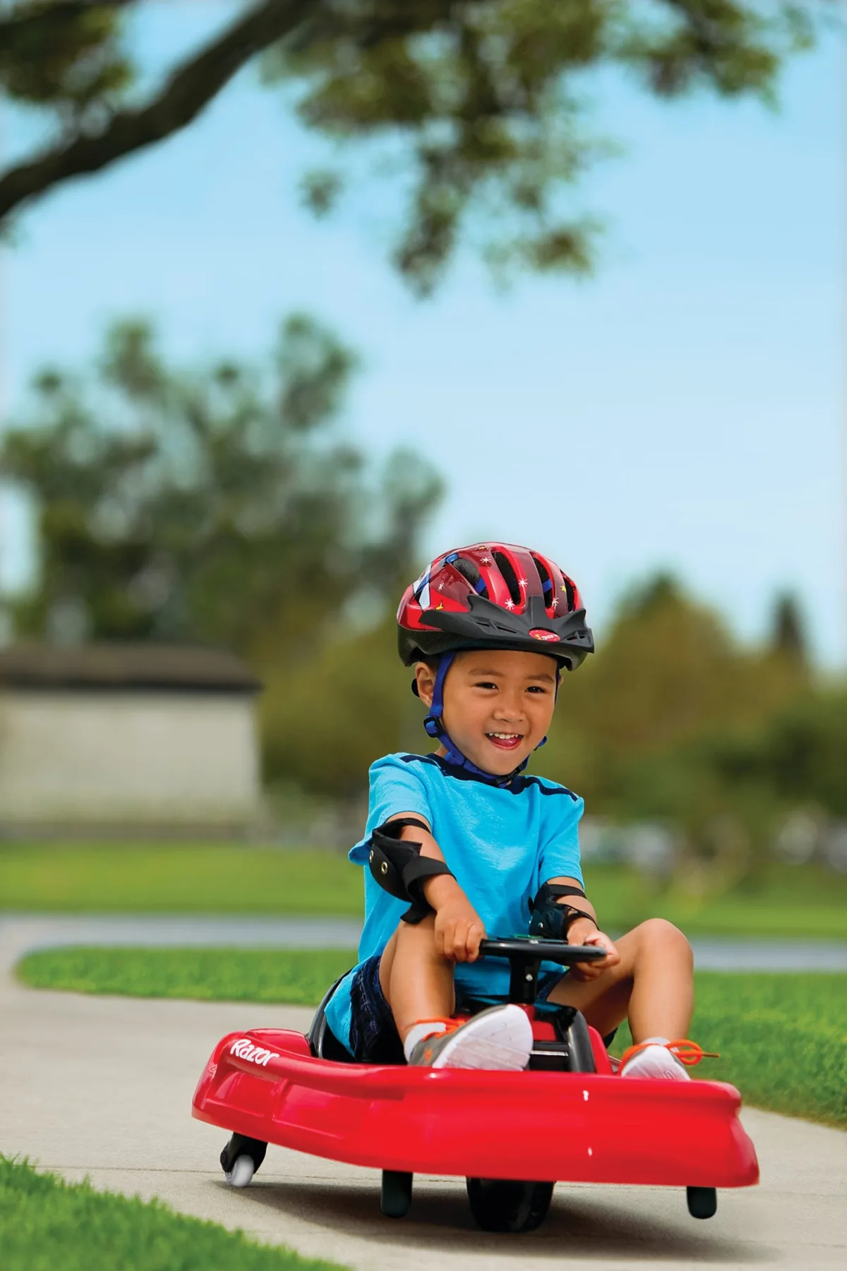 Toddler boy in helmet steering the red Razor Lil Crazy electric ride-on along a garden path with trees in background