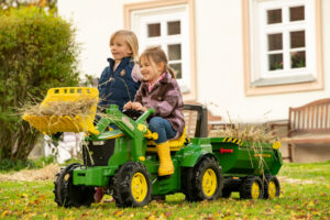 A child riding the large green Rolly Toys John Deere 7310R pedal tractor, which includes a working front loader and a large matching dumper trailer.